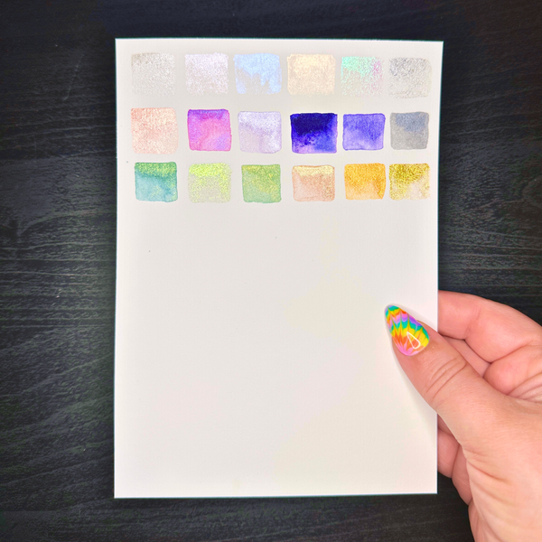 Sheet of watercolor swatches held by a hand with colorful nail polish on a dark surface.