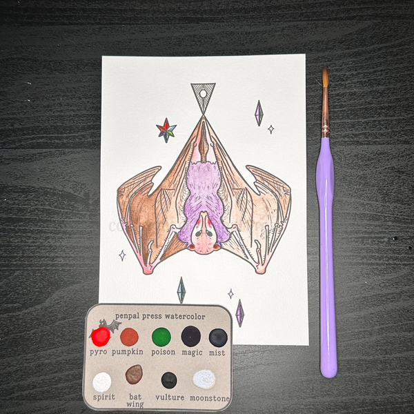 A watercolor painting of a hanging bat on white paper, shown with a purple-handled paintbrush and a small watercolor palette on a dark wood surface. The palette is labeled with colors including pyro pumpkin, poison magic mist, spirit, bat wing, and vulture moonstone.