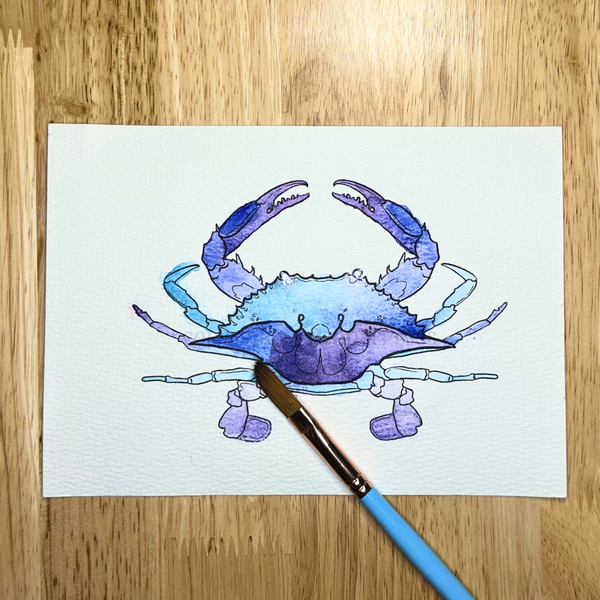 Watercolor painting of a blue crab on white paper with a paintbrush on a wooden surface.
