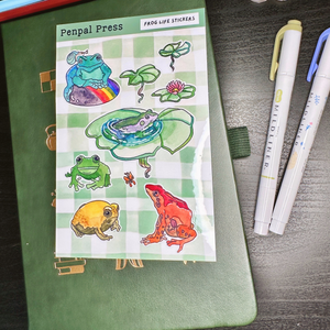 Sheet of frog-themed stickers by Penpal Press on a green notebook with pens beside it.