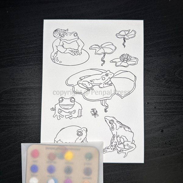 A linework design of different species of frogs with lily pads. a snail, a small butterfly, a rock. Shown with a set of watercolor samples in a rainbow of colors, sitting on a dark wood background.
