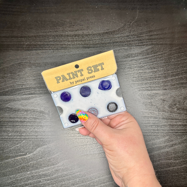 Hand holding a paint set with blue, black, and purple paint on a wooden surface