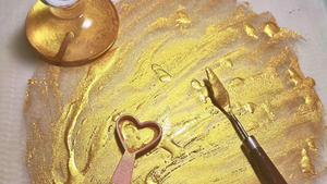 Gold paint with a pink heart-shaped measuring spoon and silver palette knife on a glass surface.