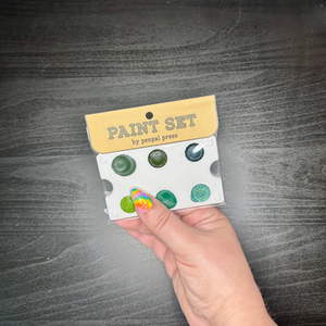 Hand holding a paint set with green and blue colors against a dark wooden surface