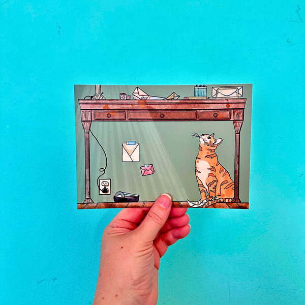 Hand holding a card with a cat sitting under a desk against a green background