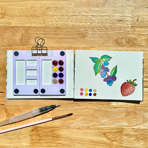 Open art supply set with watercolor paints and a drawing of blueberries and strawberry on a wooden surface.
