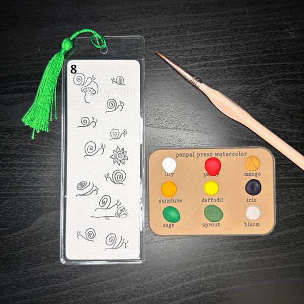 Paintable bookmark with snail designs all over, next to watercolor sample set with yellows, oranges, red, and greens, also some purple. Bookmark paper sits on top of a plastic bookmark cover and green tassel. 