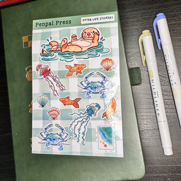 Sheet of ocean-themed stickers by Penpal Press on a green notebook with pens beside it.