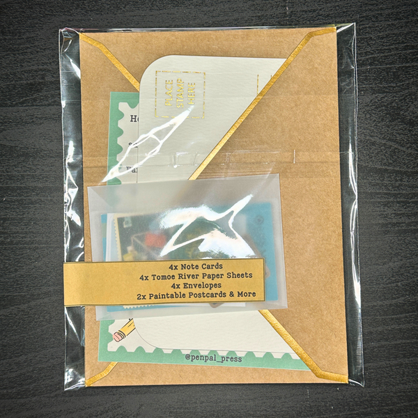 Packaging of stationery items including note cards, paper sheets, envelopes, and paintable postcards on a dark surface.