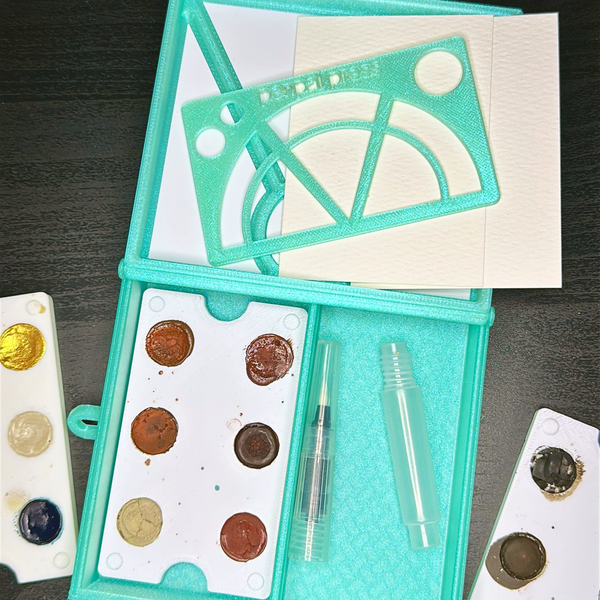 Iridescent Paint Box with two bottom divided sides holding paint sets and a travel watercolor brush. The lid is shaped like an envelope on the inside. A stencil sits on top of the box lid.