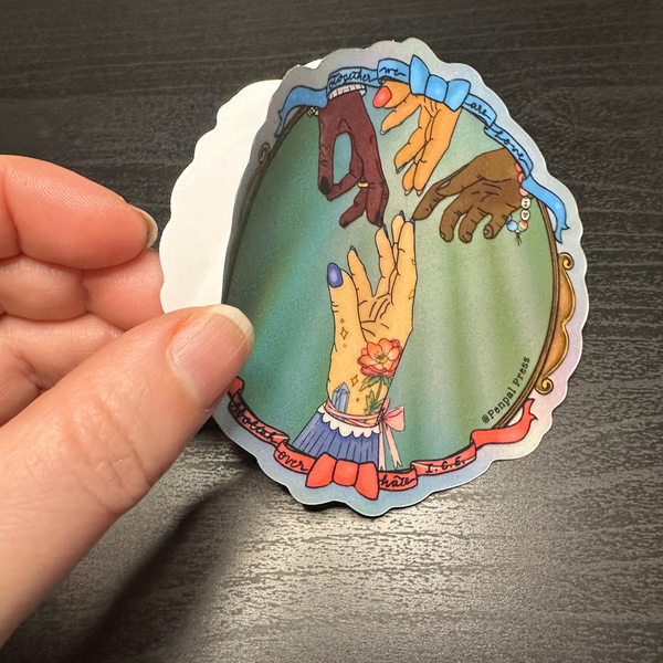 Hand holding a colorful sticker with artistic depiction of hands with a mirror finish on a dark surface