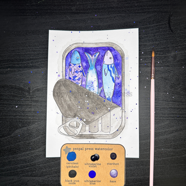 A painted design of three sardines in a metal tin using blues, purples, black/grey, and shimmery watercolors. The page is on a dark wood background with a paintbrush and set of six samples of watercolor. 