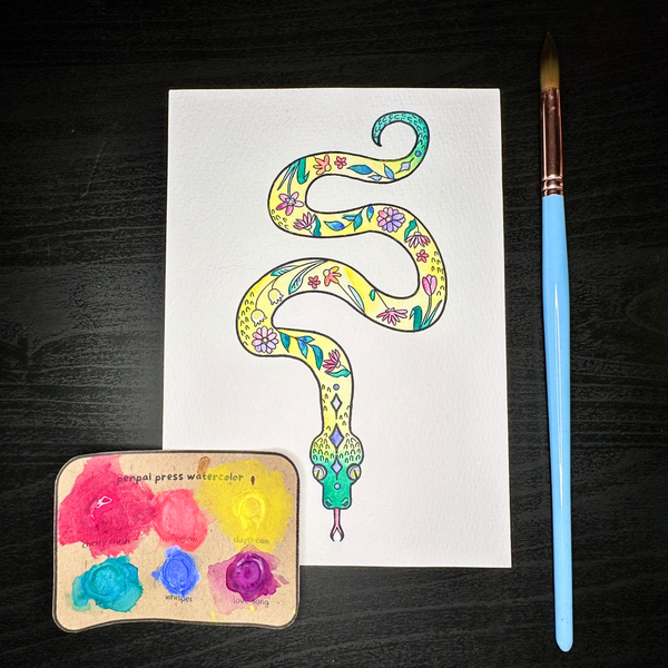 Colorful snake illustration on white paper with a paintbrush and watercolor palette on a dark surface.