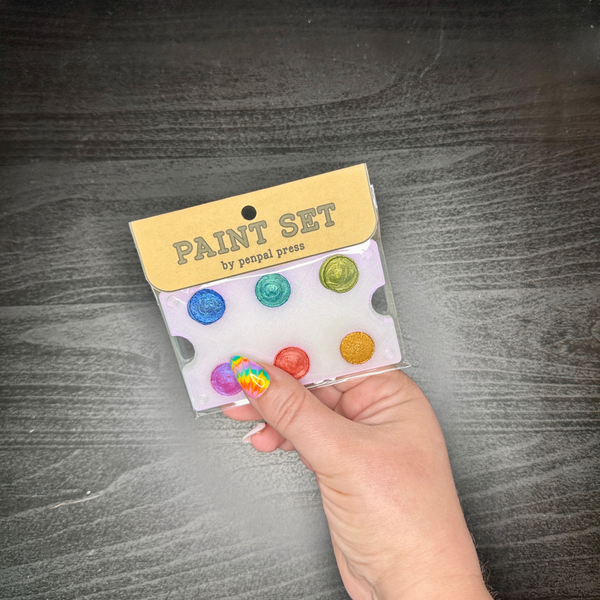 Hand holding a small paint set with colorful dots against a dark wooden surface