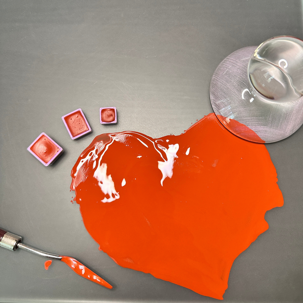 Three small watercolor pans of different sizes  filled with orange-red paint, a metal spatula with wet watercolor paint, a heart shaped area of wet venetian red paint on a glass slab, with a glass muller at the top right.