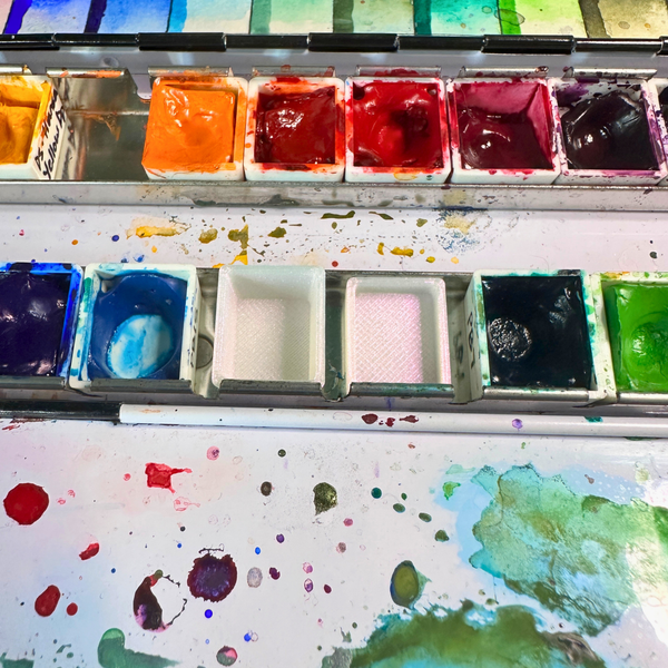 A close-up overhead view of a watercolor paint palette with multiple wells of mixed paint in red, orange, yellow, green, blue, black, and white, with paint splattered across the surface below.