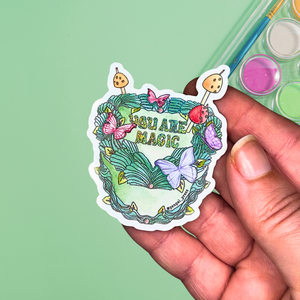 Hand holding a colorful sticker with 'You Are Magic' text against a green background of a fancy cake with butterflies and mushrooms.