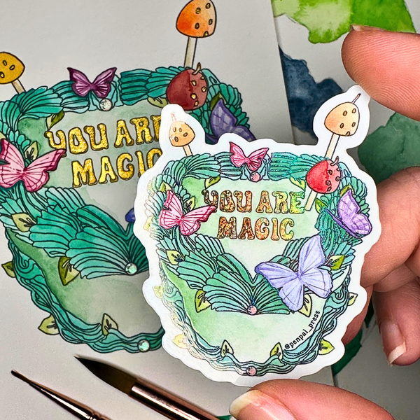 Colorful cake sticker with 'You Are Magic' text held by a hand, with a painting of it in the background.