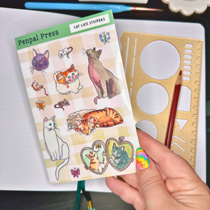 Sheet of cat-themed stickers held by a hand with a ruler and pencil in the background