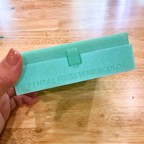 Green box with 'Penpal Press Watercolor' text held by a hand on a wooden surface
