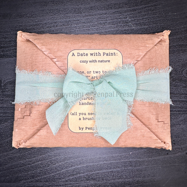 A square package wrapped in textured kraft paper, tied with a frayed sage green ribbon and bow, with a vintage-style paper label in the center, photographed from above on a dark wood background.
