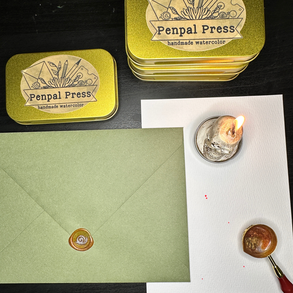 A styled flat-lay of Penpal Press handmade watercolor products on a dark tabletop, including two gold metal tins with the brand label, a sage green envelope with a wax seal, a white sheet of paper, a melting candle in a glass dish, and a wax seal stamp.