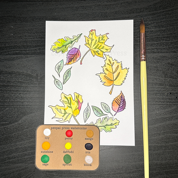 A fall leaf design painted with yellow, orange, purple, red, and green watercolor, on a dark wood background with a set of dots of watercolor and yellow handled paintbrush.
