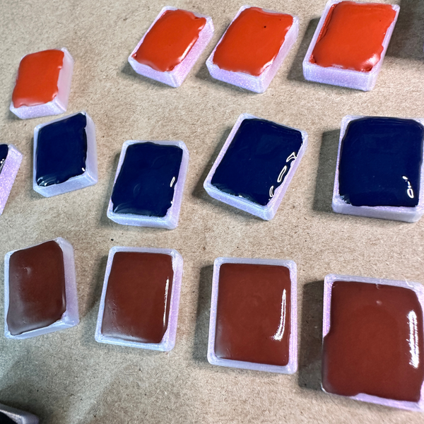 Newly filled quarter pans of watercolor paint in venetian red, Prussian blue, and violet hematite. Pans are laid out in rows on top of chipboard.