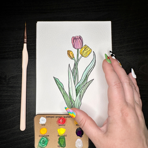 Hand holding a drawing of tulips with a paintbrush and color swatches on a dark surface