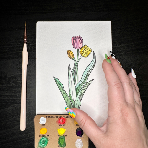 Hand holding a drawing of tulips with a paintbrush and color swatches on a dark surface