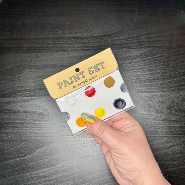 Hand holding a paint set with yellows, orange, violet, and more.