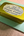 A small rectangular gold metal tin labeled 'Penpal Press handmade watercolor' sits on a light green paper envelope over a wood surface. A paintbrush is partially visible at the top left.