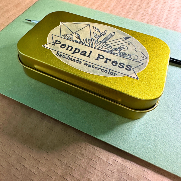 A small rectangular gold metal tin labeled 'Penpal Press handmade watercolor' sits on a light green paper envelope over a wood surface. A paintbrush is partially visible at the top left.