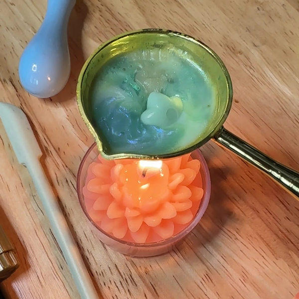 small pink candle melting green wax in a metal spoon, on a wooden surface with a spoon and other items.