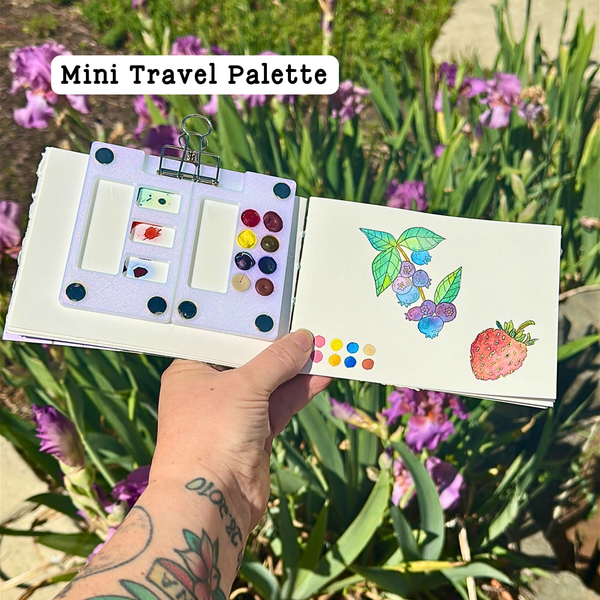 Mini travel palette with watercolor samples held in front of a garden background
