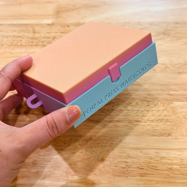 Colorful watercolor paint box with 'Penpal Press Watercolor' branding held by a hand on a wooden surface.