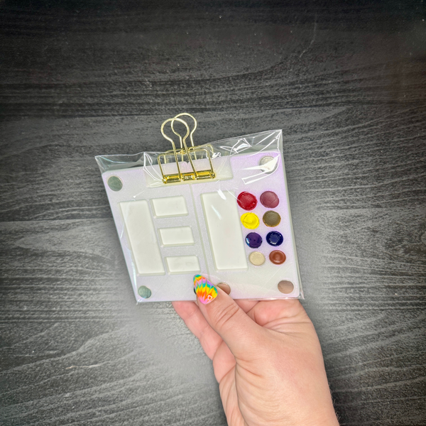 Packaged stationery items including colorful watercolor paint and a clip held by a hand on a dark surface.