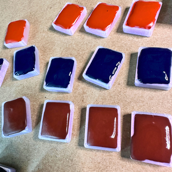 A collection of rectangular paint pains with glossy wet handmade paint in various colors including orange, dark blue, and reddish-brown, arranged on a textured kraft surface.