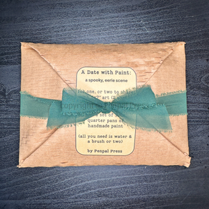 A handmade paper envelope-style package wrapped with a deep green ribbon, featuring a vintage-style label that reads 'A Date with Paint: a spooky, eerie scene' and 'by Penpal Press,' placed on a dark wood background.