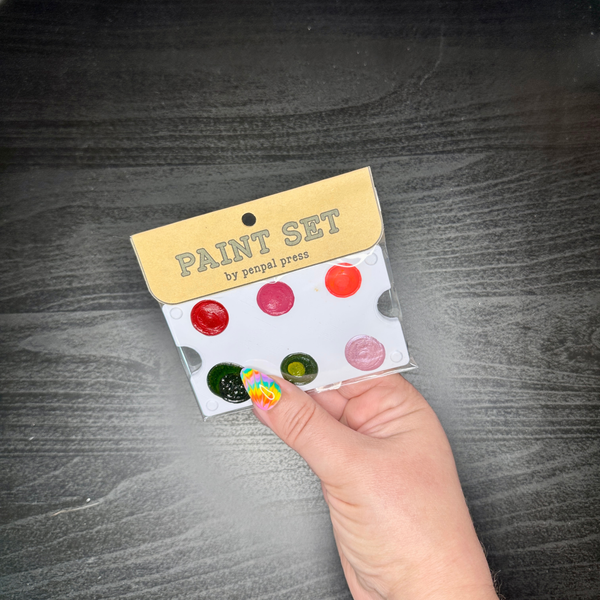 Hand holding a small paint set with colorful dots on a dark wooden surface