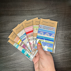 Hand holding a collection of Penpal Press washi tape samples on a wooden surface