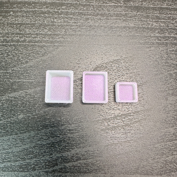 Three small white plastic pans for watercolor, arranged by size on a dark gray wood-grain surface.